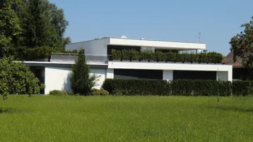 Modernes weißes Gebäude mit Flachdach und gepflegtem Rasen, umgeben von grünen Hecken.