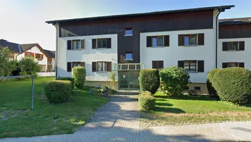 Mehrstöckiges Wohnhaus mit weißer Fassade, braunen Holzverkleidungen und Fensterläden, umgeben von einem gepflegten Vorgarten.