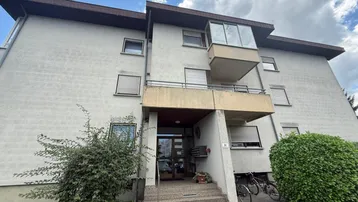 Mehrfamilienhaus mit mehreren Etagen, Balkonen und Jalousien an den Fenstern unter bewölktem Himmel.