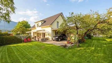 Haus mit großem Garten, Terrasse und Carport unter blauem Himmel und Bergblick.