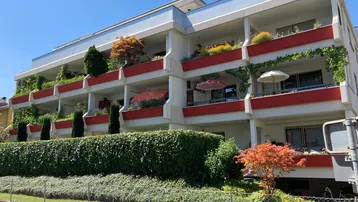 Mehrfamilienhaus mit mehreren Balkonen, die mit Pflanzen und Sonnenschirmen ausgestattet sind, umgeben von gepflegter Hecke.