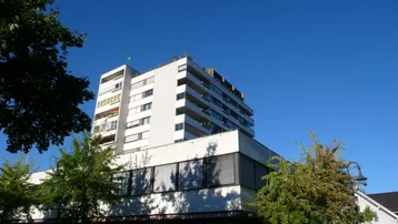 Modernes Mehrfamilienhaus mit mehreren Balkonen und einer Dachterrasse unter blauem Himmel.