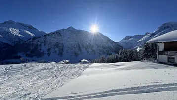 Sonnige Winterlandschaft mit schneebedeckten Bergen, Skipisten und einem traditionellen Gebäude im Alpenstil.
