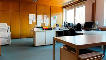 Geräumiges Büro mit Holzdeckenverkleidung, Teppichboden und großen Fenstern, die viel Tageslicht hereinlassen.