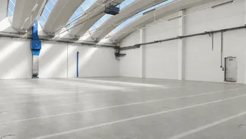 Große, helle Lagerhalle mit hohen Decken, Betonboden und Oberlichtern für viel Tageslicht.