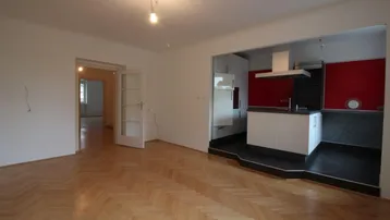 Heller Wohnbereich mit Fischgrätparkett und offener Küche mit roten Akzenten und Kochinsel.