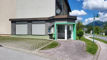 Gebäudeansicht mit Ladenfront, Rollläden und gepflastertem Vorplatz unter blauem Himmel.
