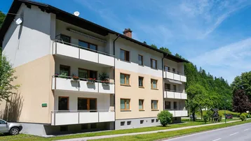 Mehrfamilienhaus mit beiger und weißer Fassade, mehreren Balkonen und Blick ins Grüne an einem sonnigen Tag.