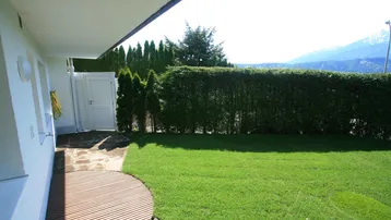 Sonniger Garten mit Rasen, hoher Hecke und Bergblick im Hintergrund.