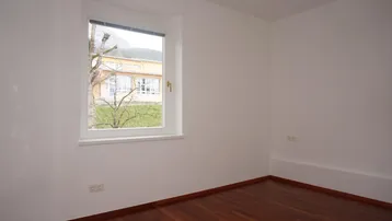Helles, leeres Zimmer mit Holzboden und Fensterblick ins Grüne.