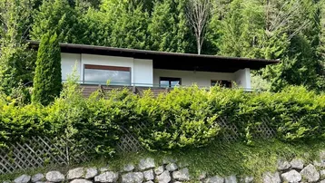 Modernes Haus mit Flachdach und großen Fenstern, eingebettet in eine grüne Berglandschaft.