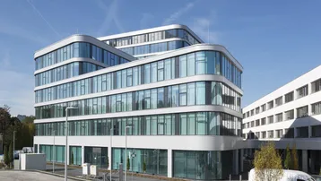 Modernes Bürogebäude mit geschwungener Glasfassade und vielen Fenstern unter blauem Himmel.