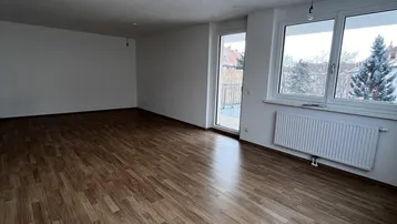 Geräumiges, leeres Wohnzimmer mit Laminatboden, großen Fenstern und Zugang zum Balkon.