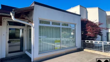 Außenansicht eines modernen Gebäudes mit großen Fenstern, weißer Fassade und einer gepflasterten Terrasse.