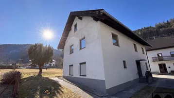 Traditionelles Wohnhaus mit weißer Fassade und Satteldach, umgeben von einer ländlichen Berglandschaft.