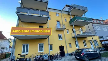 Modernes gelbes Mehrfamilienhaus mit mehreren Balkonen, großen Fenstern und Stellplätzen.