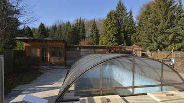 Poolbereich mit Überdachung und angrenzendem Holzhaus unter blauem Himmel.