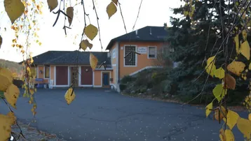 Gebäudeansicht mit Zufahrt und herbstlicher Vegetation im Vordergrund, umrahmt von gelben Blättern.