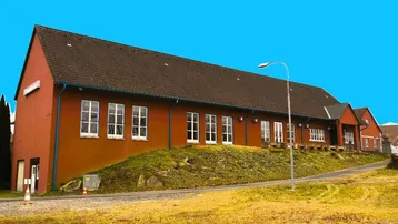Lange Seitenansicht des roten Gebäudes mit vielen Fenstern und einer leicht abfallenden Grünfläche.