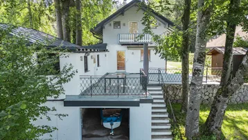 Weißes Haus mit Balkon, Terrasse und integrierter Bootsgarage, umgeben von hohen Bäumen.
