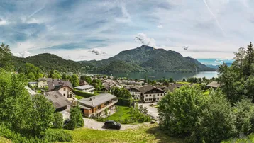 Panoramablick auf eine malerische Stadt am See, umgeben von grünen Bergen unter blauem Himmel.