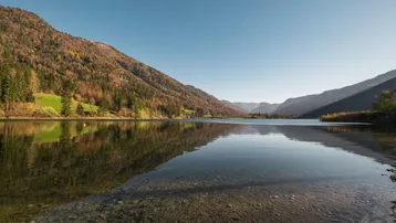 Idyllischer See umgeben von bewaldeten Bergen unter klarem Himmel, ideal für Naturliebhaber.