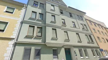 Mehrstöckiges Wohngebäude mit hellgrüner Fassade, vielen Fenstern und Rollläden in einer städtischen Umgebung.