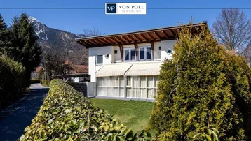 Modernes Einfamilienhaus mit gepflegtem Vorgarten und Bergblick unter blauem Himmel.