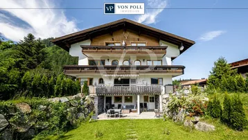 Traditionelles Alpenhaus mit mehreren Balkonen und einer Terrasse im Grünen, umgeben von Bergen.