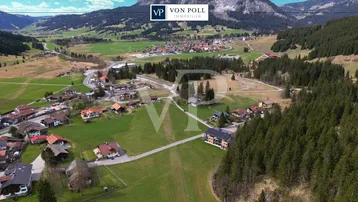 Panoramablick aus der Vogelperspektive auf ein malerisches Dorf in einer Berglandschaft mit grünen Wiesen und Wäldern.
