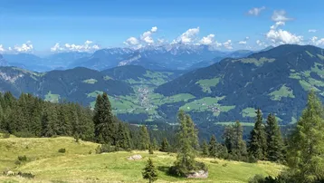 Panoramablick auf eine weite Berglandschaft mit grünen Hügeln, Wäldern und einem Tal mit einem Dorf unter blauem Himmel.