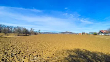 Weitläufiges Ackerland unter einem klaren blauen Himmel mit entfernten Häusern und Hügeln am Horizont.