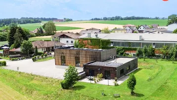 Luftaufnahme eines modernen Gebäudekomplexes mit Holzfassade, umgeben von grünen Feldern und ländlicher Bebauung.
