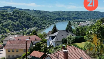 Panoramablick über eine Wohnsiedlung mit Fluss und bewaldeten Hügeln unter blauem Himmel.