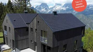 Modernes Haus mit dunkler Holzfassade, Satteldach und großer Terrasse, umgeben von einer beeindruckenden Berglandschaft.