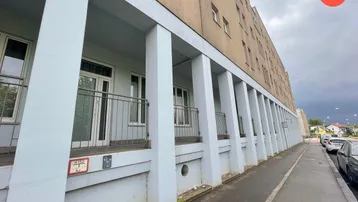 Langes Wohngebäude mit mehreren Loggien und Balkonen, davor ein Gehweg und parkende Autos unter bewölktem Himmel.