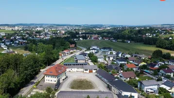 Luftaufnahme des Anwesens mit mehreren Gebäuden, umgeben von Wohnsiedlungen, Feldern und Wäldern unter blauem Himmel.