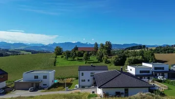 Weitläufige Luftaufnahme einer Wohnsiedlung mit modernen Häusern und grüner Hügellandschaft im Hintergrund.