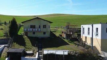 Gelbes Haus in Hanglage mit weiten grünen Feldern und einem weiteren Gebäude im Hintergrund.
