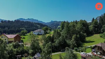 Weitläufige Landschaft mit grünen Hügeln, Bäumen und vereinzelten Häusern unter blauem Himmel.