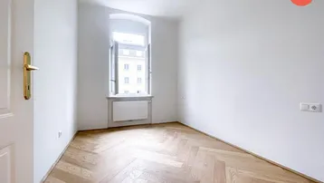 Helles Zimmer mit Fischgrätparkett, hohen Decken und einem großen Fenster für viel Tageslicht.