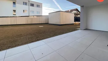 Große geflieste Terrasse mit Zugang zu einer Rasenfläche und Blick auf moderne Gebäude.
