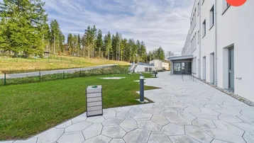 Moderne Gebäudefassade mit gepflastertem Weg, Rasenfläche und bewaldetem Hügel im Hintergrund unter blauem Himmel.
