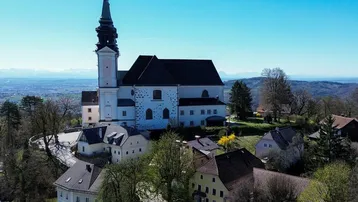 Luftaufnahme einer historischen Kirche und umliegender Gebäude auf einem Hügel mit weitem Blick auf die Landschaft und Berge.