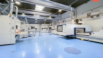 Großer, heller Industriebereich mit blauer Bodenfläche und modernen Maschinen unter sichtbaren Lüftungsrohren.