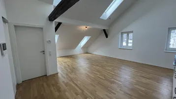Heller Wohnbereich mit Holzbalken, Dachschrägen und Parkettboden, beleuchtet durch Dachfenster.
