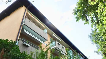 Gebäudeansicht mit mehreren Balkonen und Markisen, umgeben von grünen Bäumen unter blauem Himmel.