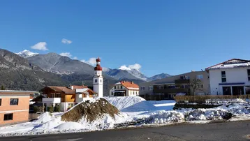 Panoramablick auf eine verschneite Wohnsiedlung mit Kirche und majestätischen Bergen im Hintergrund unter blauem Himmel.