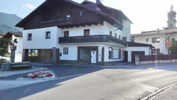 Traditionelles Gebäude mit weißer Fassade, Holzbalkonen und einem Ladenlokal im Erdgeschoss, umgeben von einer ländlichen Umgebung.