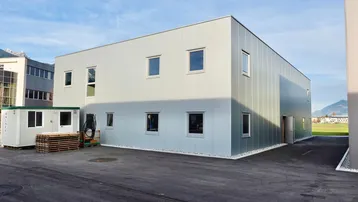 Modernes Geschäftsgebäude mit heller Fassade und asphaltiertem Vorplatz unter blauem Himmel.
